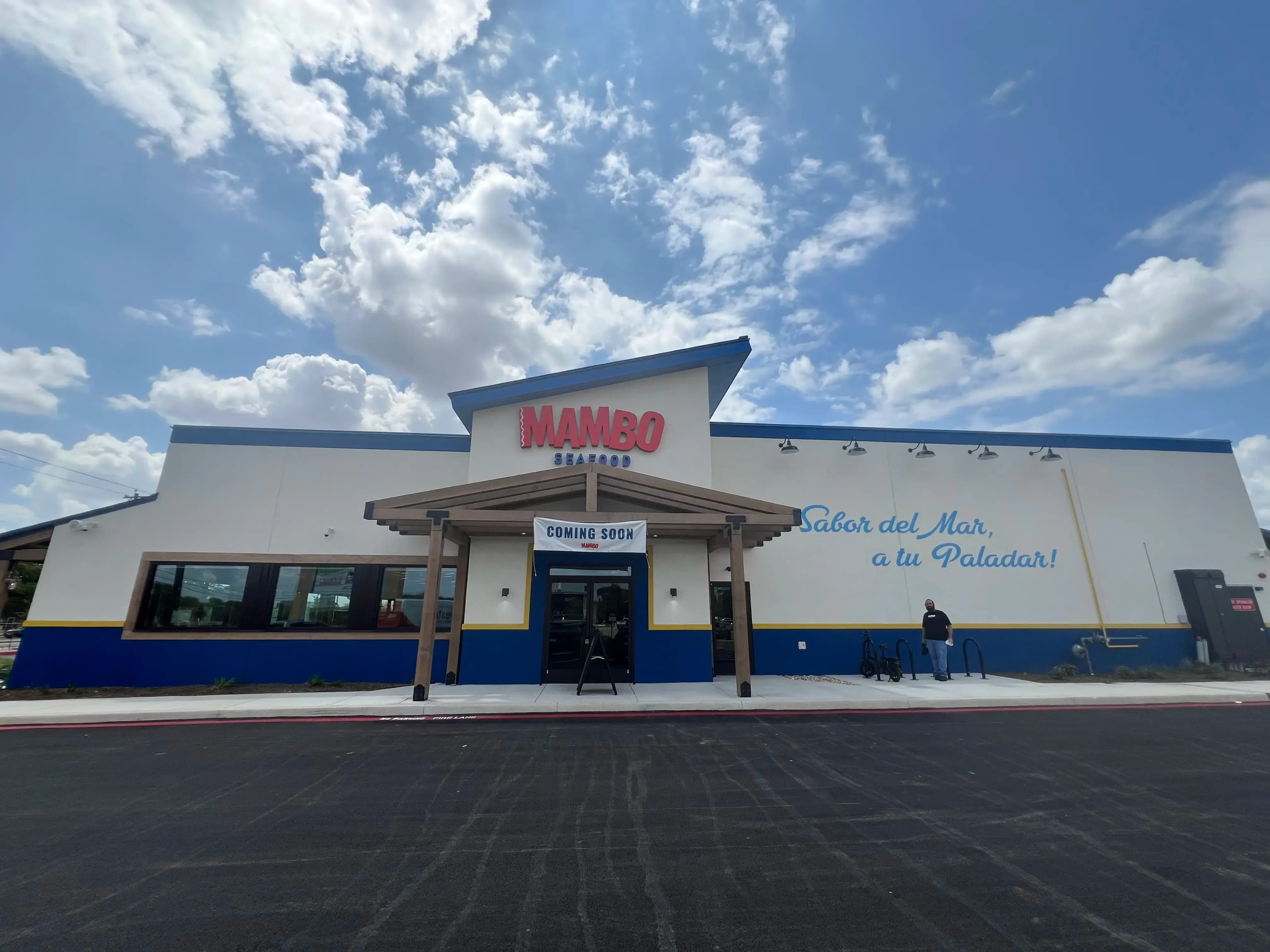 A wide-angle daytime shot of a Mambo Seafood restaurant with a modern, white and blue exterior. A "Coming Soon" banner hangs over the wooden-framed entrance under a bright, cloudy sky. A Spanish slogan, "Sabor del Mar, a tu Paladar!" is painted on the right wall.