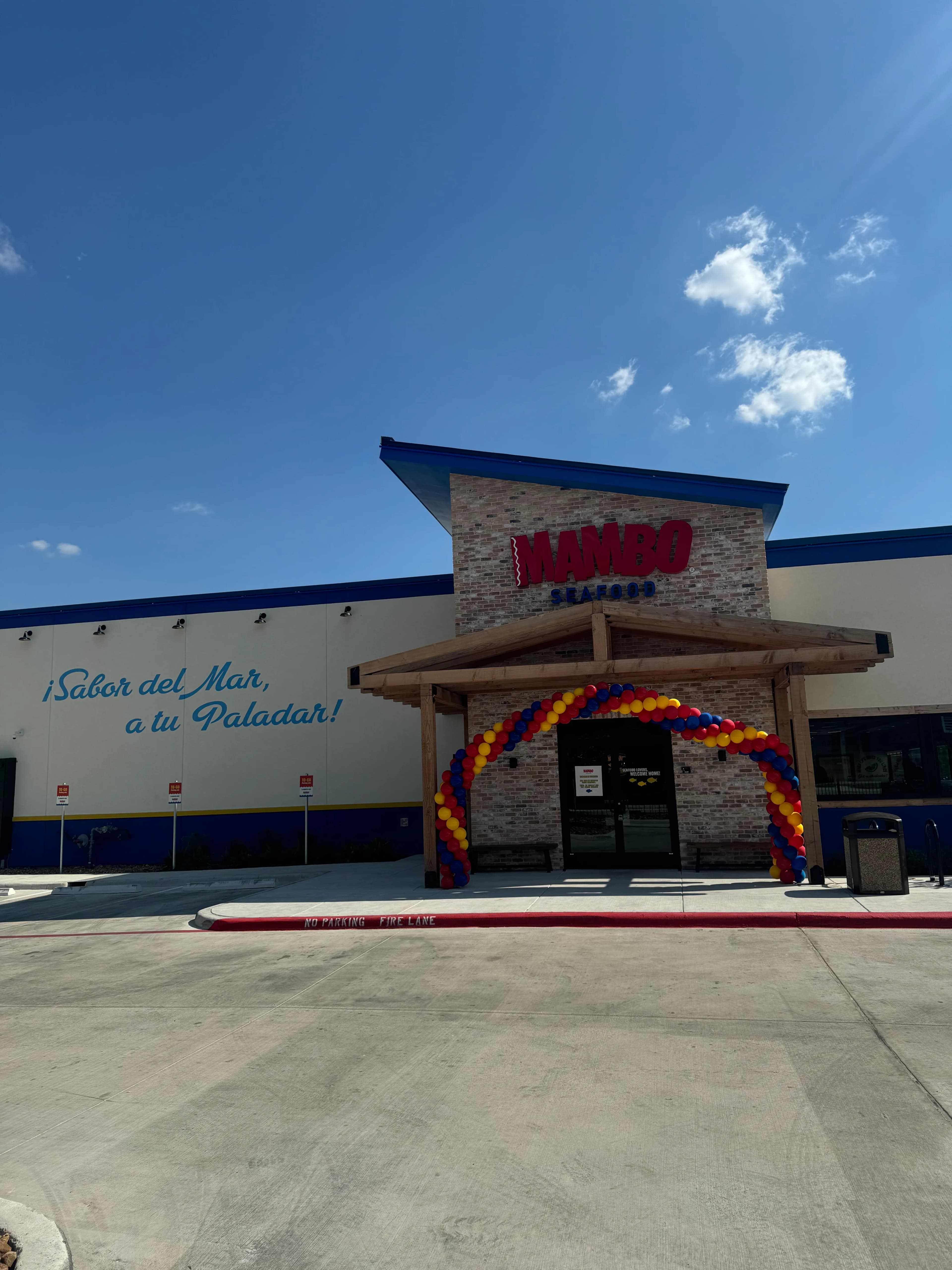 A Mambo Seafood restaurant exterior on a sunny day, featuring a brick entrance decorated with a festive red, yellow, and blue balloon arch. The side of the building is white with blue trim and a painted Spanish slogan.