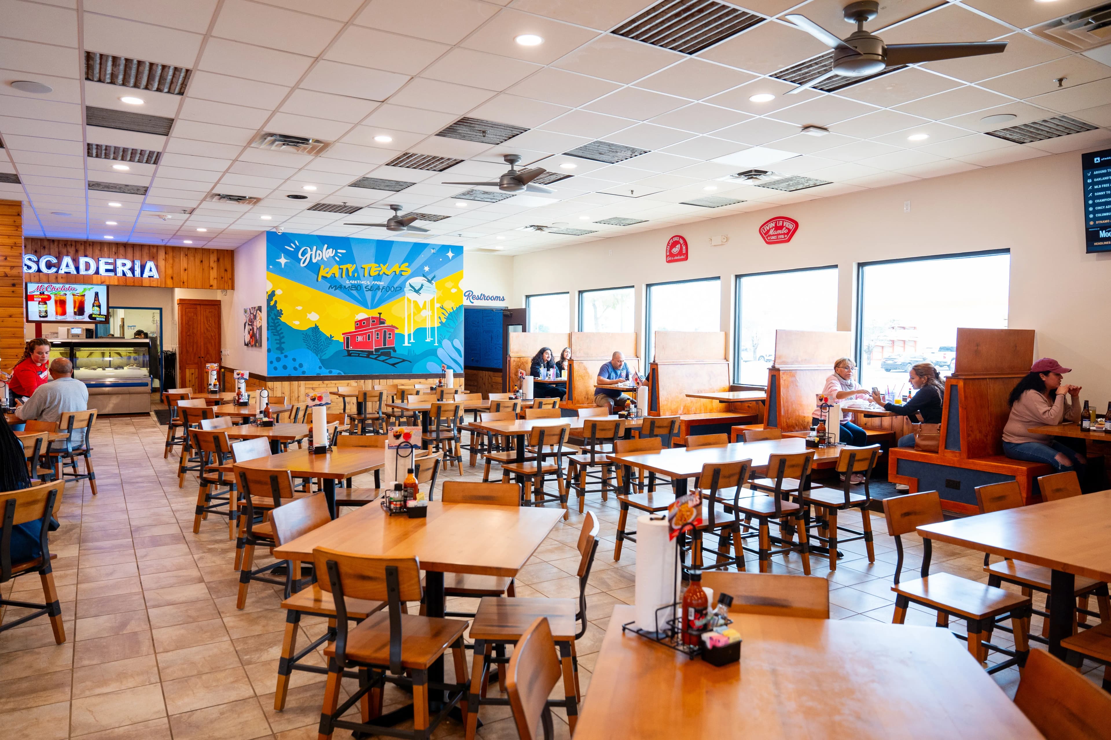 The interior of a bright, spacious restaurant with many wooden tables and chairs. A large, colorful mural on a back wall says "Hola Katy, Texas" and depicts a red train car. A sign in the background marks the "Pescaderia" (fish market) section.