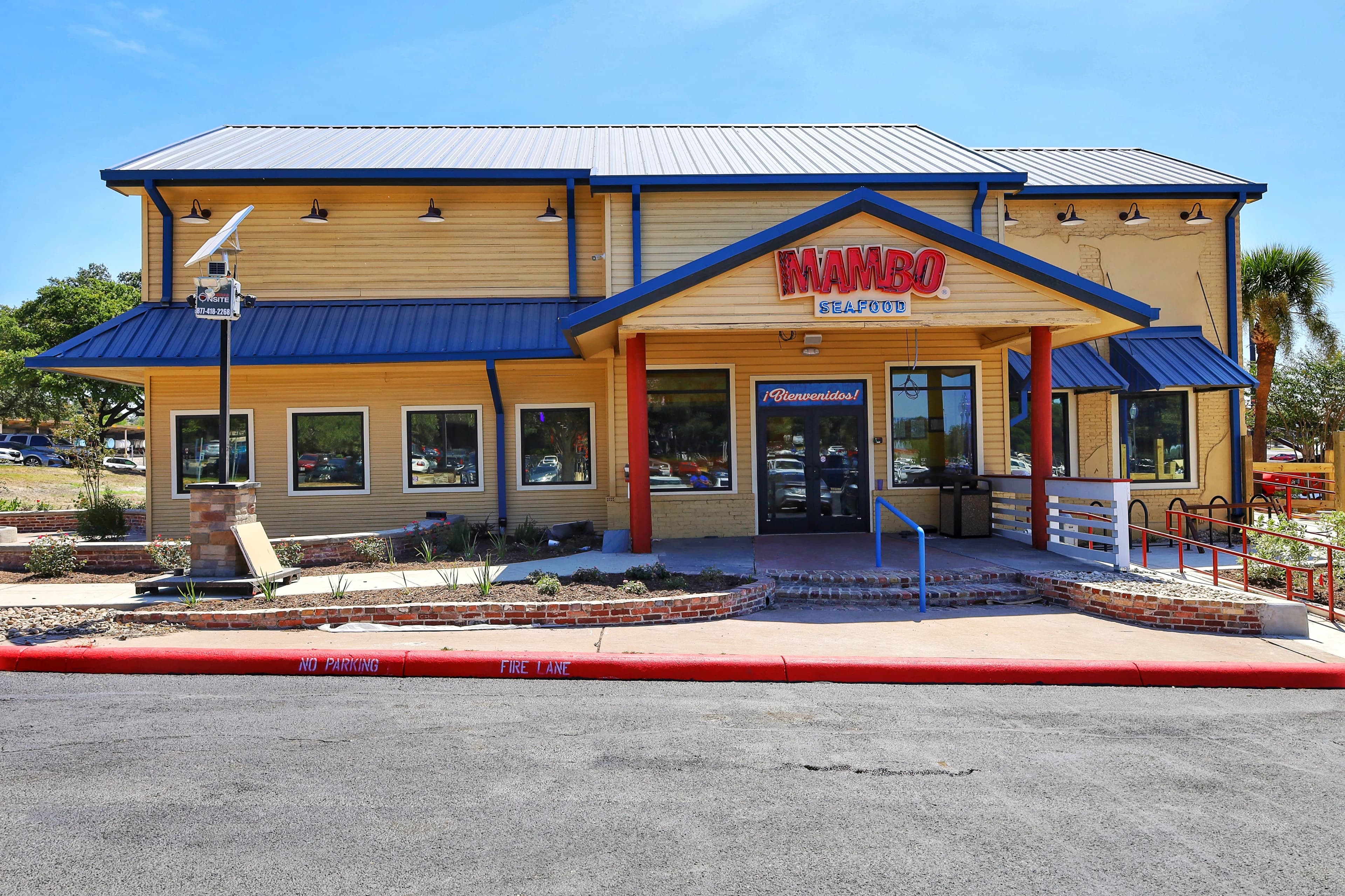 A two-story restaurant building with yellow horizontal siding and a blue metal roof. The Mambo Seafood logo is centered over a covered entrance supported by red pillars, and a "No Parking Fire Lane" is painted on the curb in the foreground.