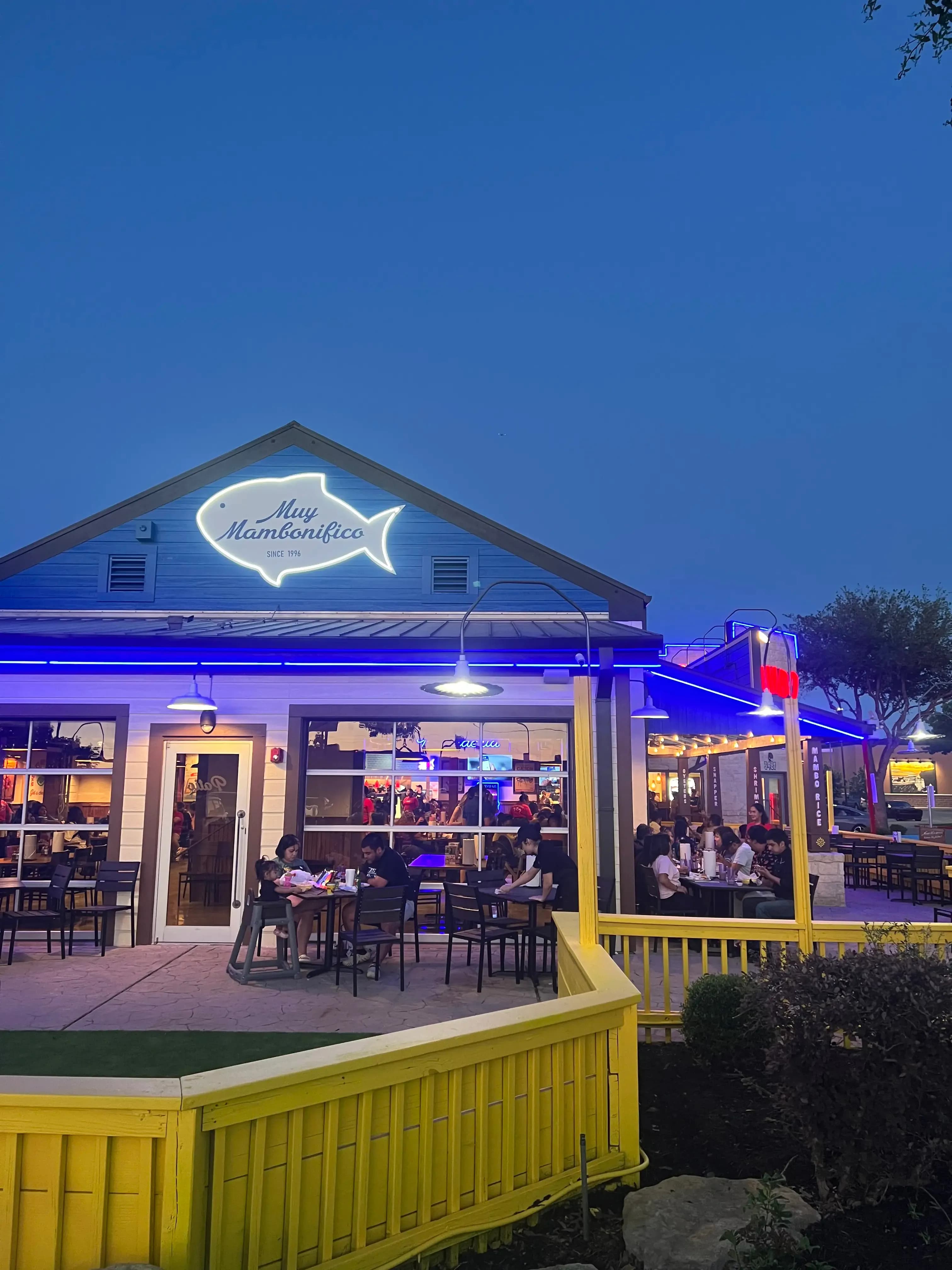 A night shot of a restaurant patio featuring a large neon sign in the shape of a fish that reads "Muy Mambonifico." People are seated at outdoor tables under blue accent lighting and warm patio lamps, separated from the foreground by a bright yellow wooden fence.